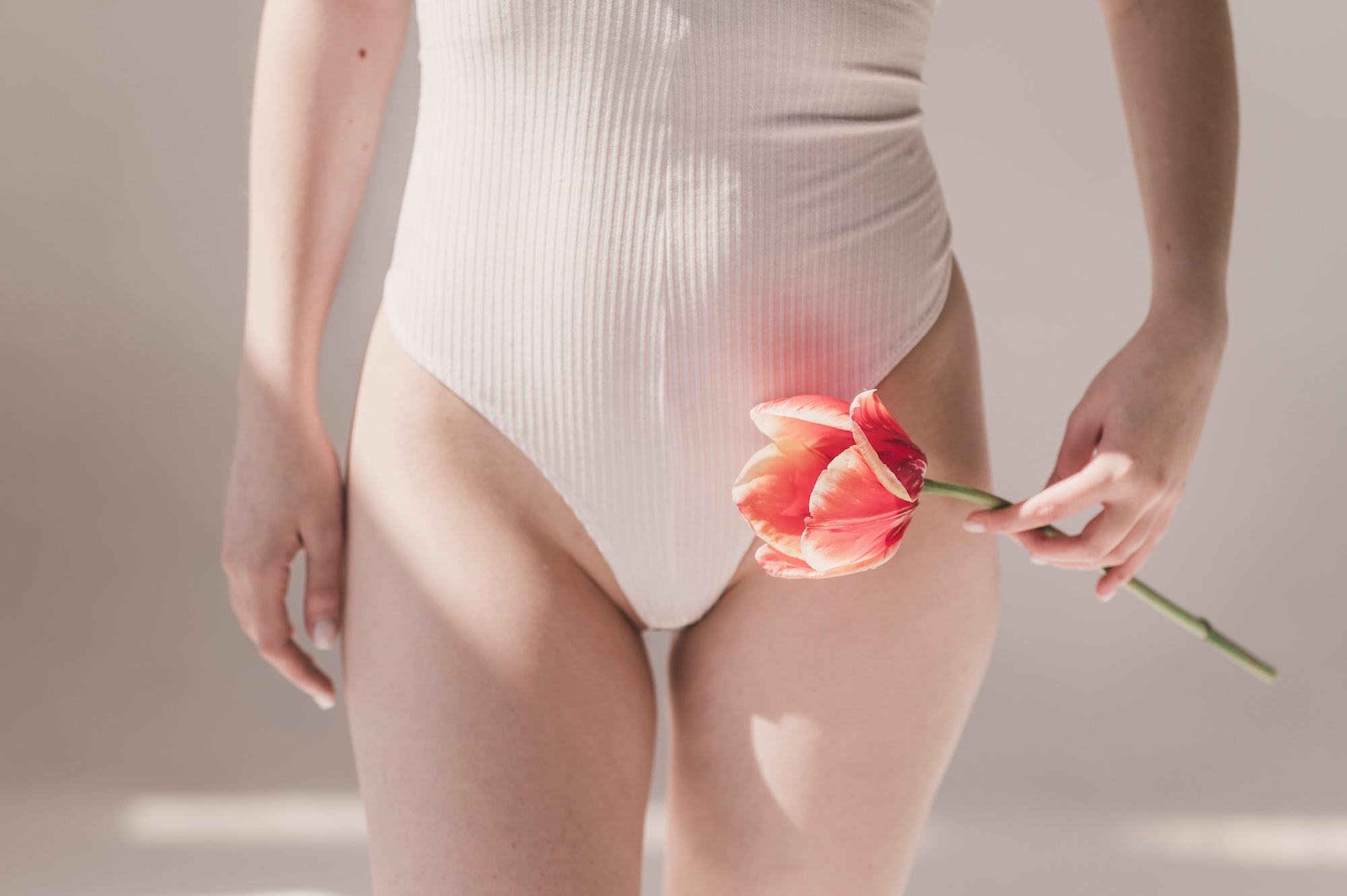 Woman in white ribbed bodysuit holding pink tulip near pelvic area, soft natural light, feminine health concept