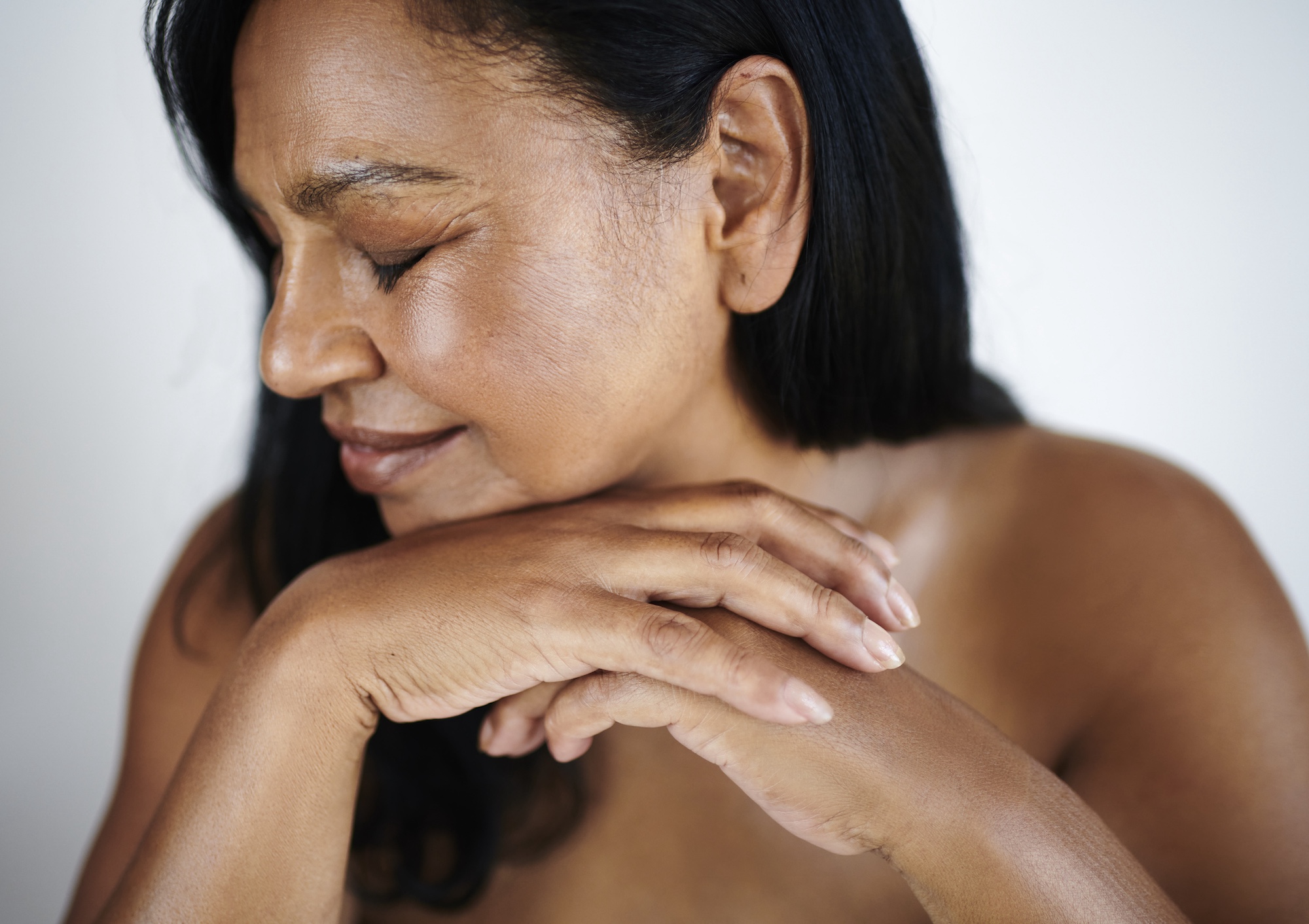 Close-up of a woman with eyes closed, resting her chin on folded hands, showcasing natural glowing skin.