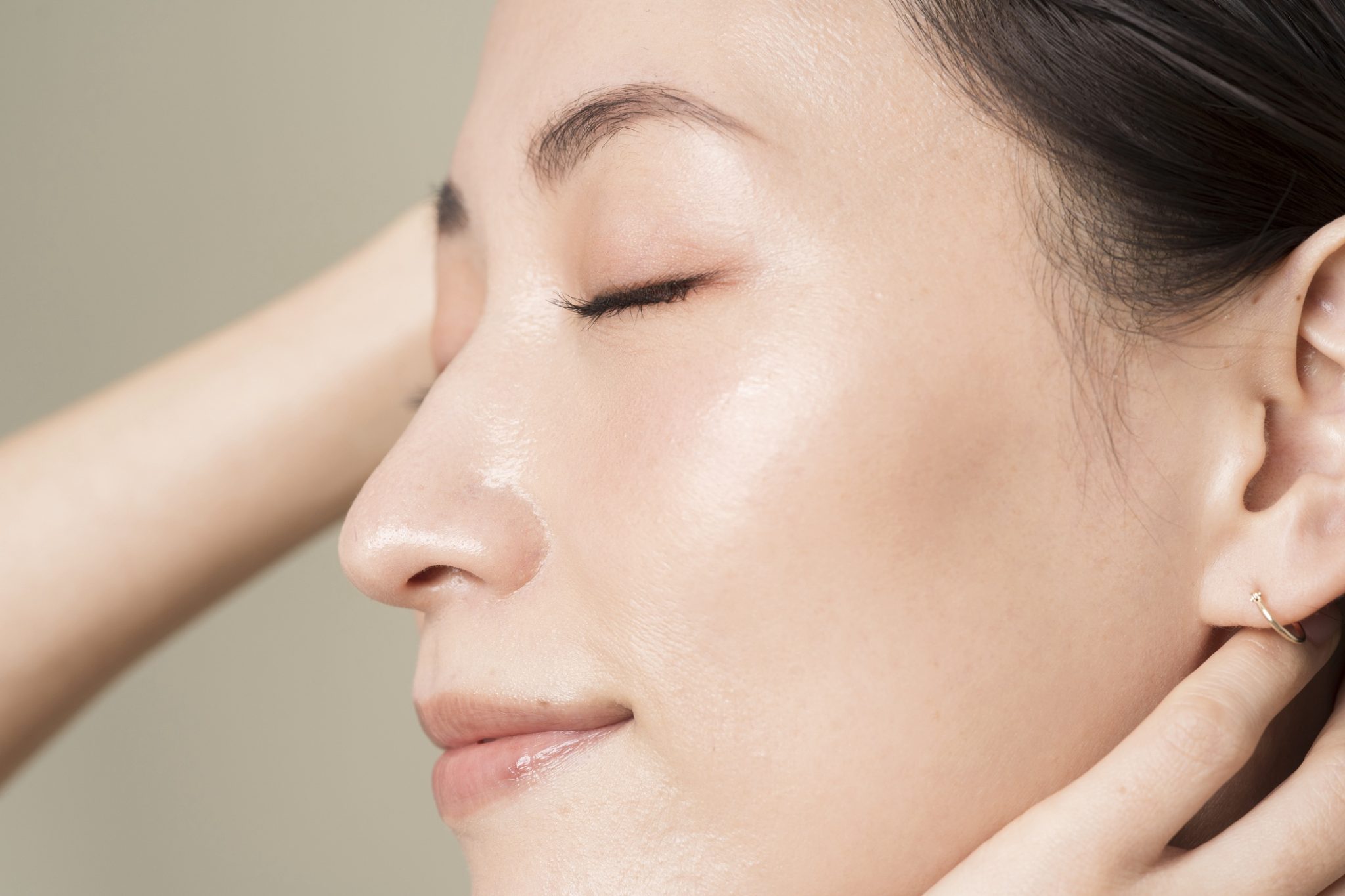 Close-up of a woman with glowing skin and closed eyes, touching her ear showcasing smooth complexion and natural makeup.