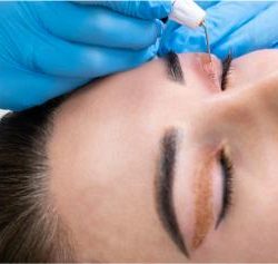 Woman receiving permanent eyeliner tattoo along upper lash line from a gloved technician.